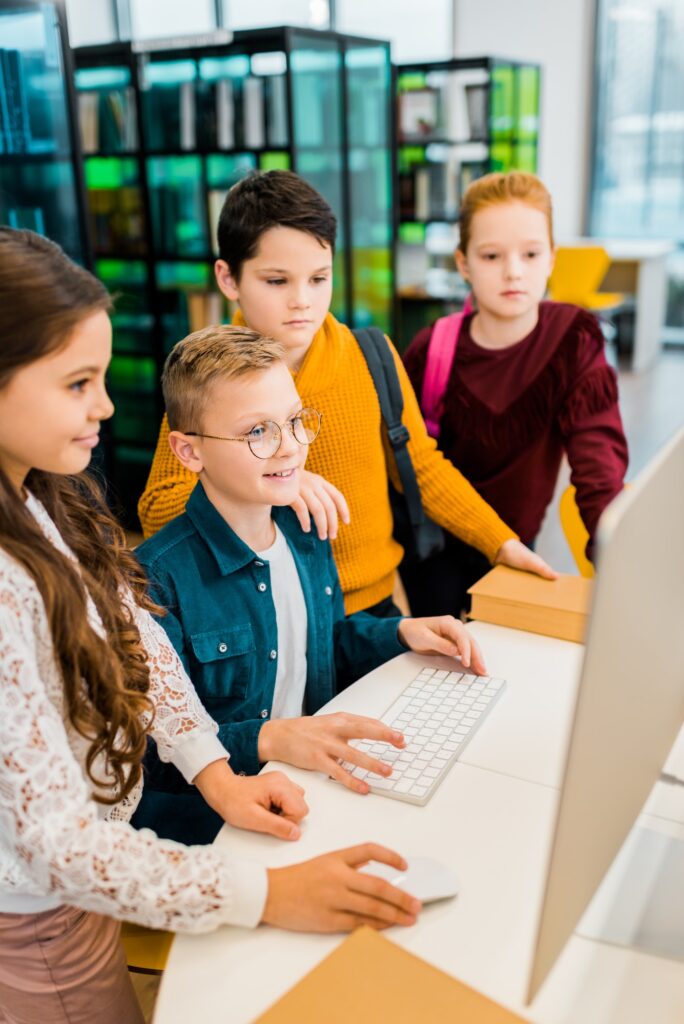 high angle view of adorable smiling schoolkids using desktop computer in library